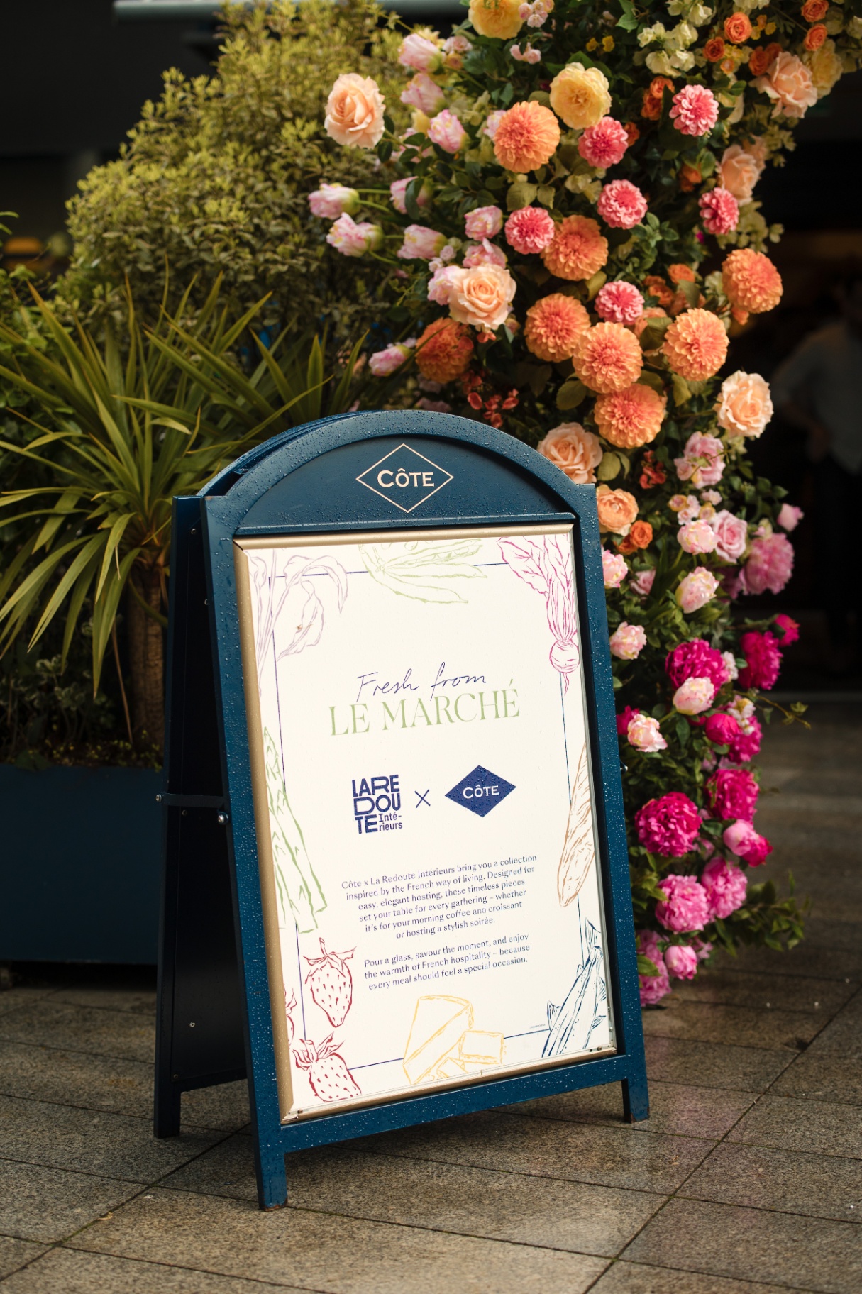 Côte A-board outside a restaurant with the 'Fresh from Le Marché' poster and Côte x La Redoute Intérieurs logos, framed by flowers.