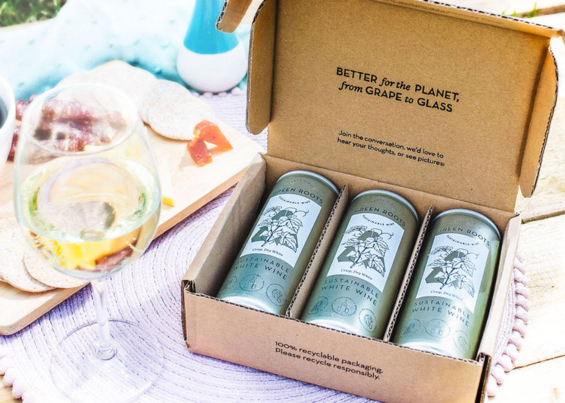 Open Green Roots box of three sustainable white wine cans beside a glass of white wine.
