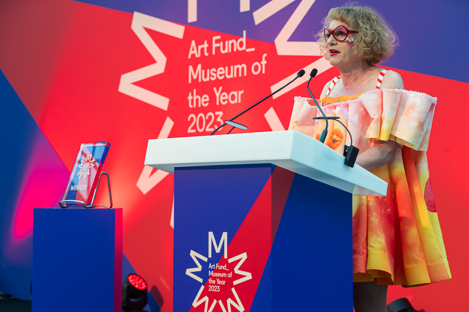 Grayson Perry speaking at the Museum of the Year 2023 ceremony in front of a red and blue starburst backdrop.