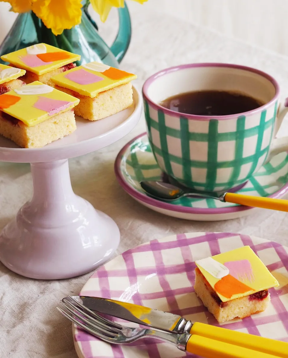 Close-up of Happy Hour Cakes petit fours on a checked plate beside a cup of coffee.