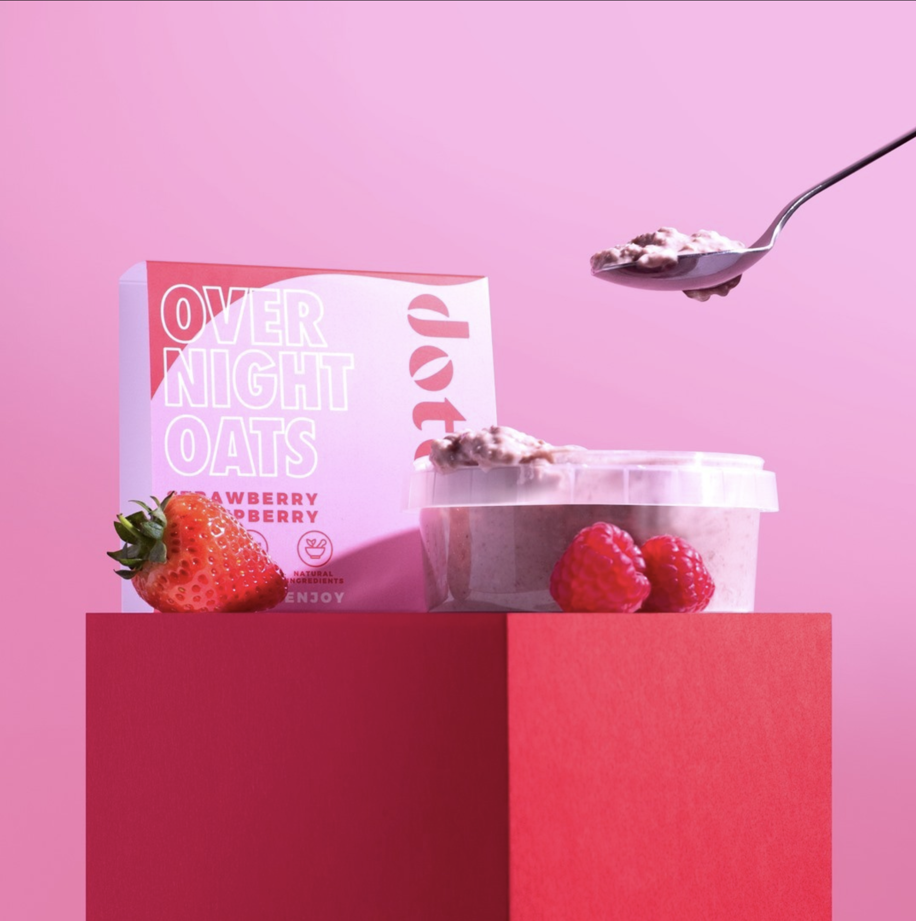 Dote strawberry and raspberry overnight oats carton and pot on a red plinth with fresh berries and a spoonful of oats.